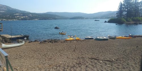 A sandy beach by a lake with kayaks and boats lined up, surrounded by mountains and trees under a clear sky.
