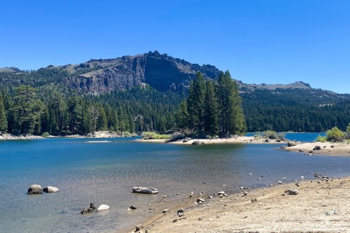 A serene lake surrounded by trees and mountains under a clear blue sky, with sandy shores and scattered rocks.