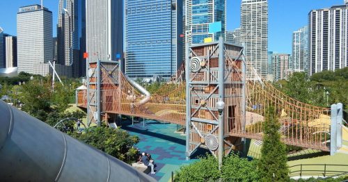 A playground with a bridge and slide, surrounded by skyscrapers and greenery in a city park on a clear day.