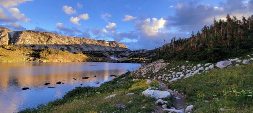 Scenic landscape of a lake surrounded by mountains and trees, with a colorful sky and rocky shoreline.