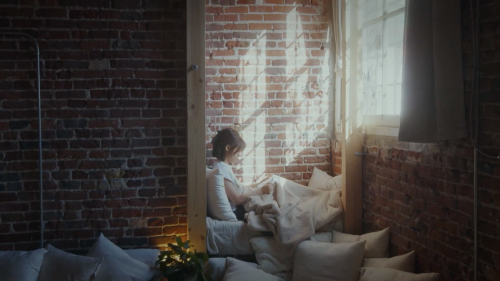 A child sits on a pile of pillows in a cozy room with exposed brick walls, reading in soft, natural light.