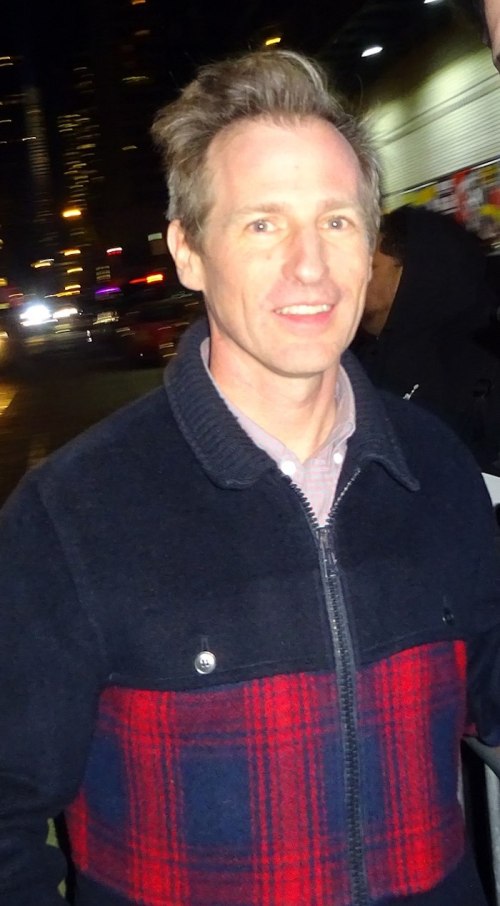 A man with light hair smiles while wearing a dark jacket with a red plaid pattern, set against a city night backdrop.