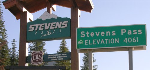 Sign for Stevens Pass with elevation 4061, surrounded by trees and mountains in a clear blue sky.