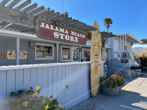 A beach store with a sign reading "Jalama Beach Store," featuring a surfboard and colorful signage.