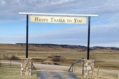 A sign reading "Happy Trails to You" stands at the entrance to a scenic, open landscape with rolling hills.