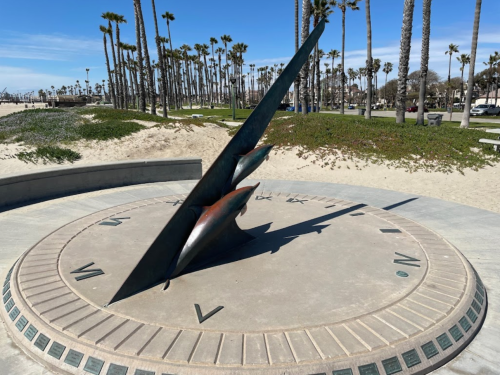 A large metal sundial on a circular base, surrounded by palm trees and a sandy beach under a clear blue sky.