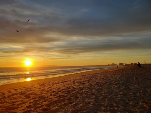 A serene beach at sunset, with gentle waves, a sandy shore, and a person biking in the distance under a colorful sky.