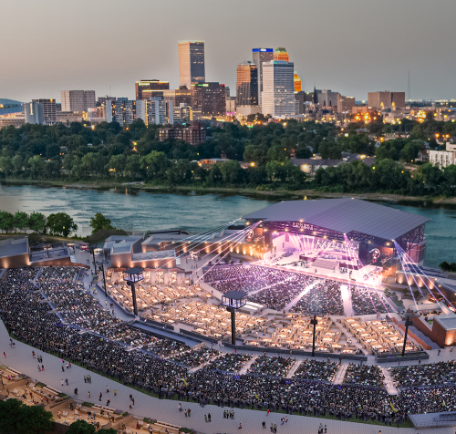 Aerial view of a large outdoor concert venue filled with people, with a city skyline and river in the background at dusk.