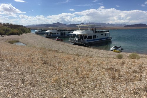 Three houseboats docked along a pebbly shore with mountains and a cloudy sky in the background.