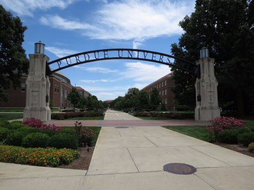 Archway sign reading "Purdue University" with a pathway lined by trees and flowers under a blue sky.
