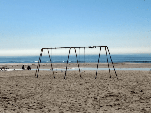 A swing set on a sandy beach with calm ocean waves and a clear blue sky in the background.