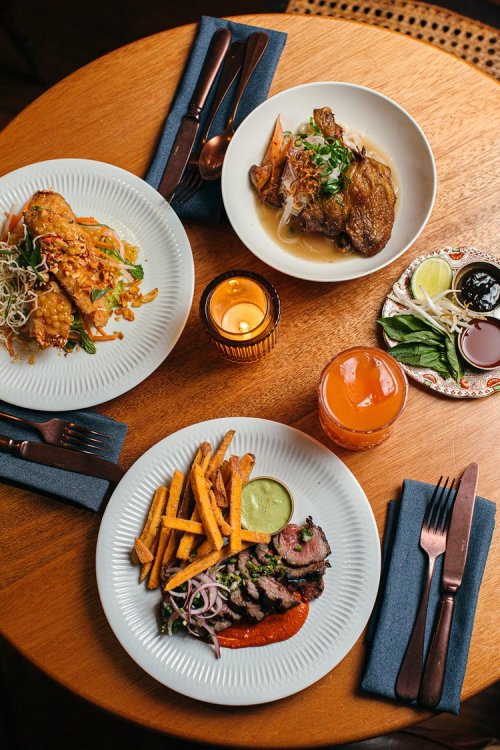 A wooden table with three dishes: fried tacos, duck with vegetables, and grilled meat with fries and sauces.