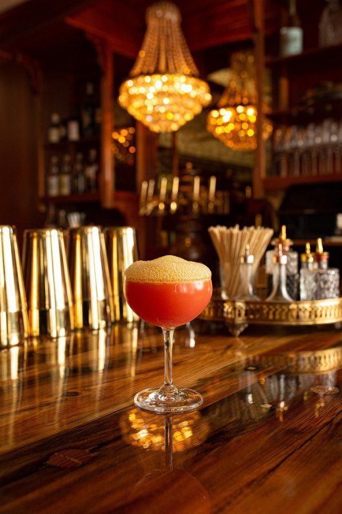 A vibrant cocktail with a frothy top sits on a polished wooden bar, illuminated by sparkling chandeliers in the background.