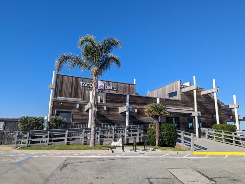 A Taco Bell restaurant with a wooden exterior and palm tree, set against a clear blue sky.