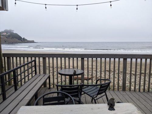 A coastal view from a deck with tables and chairs, overlooking a foggy beach and calm ocean waves.