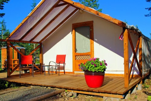 A cozy tent cabin with a wooden deck, two red chairs, and a potted flower on a sunny day.