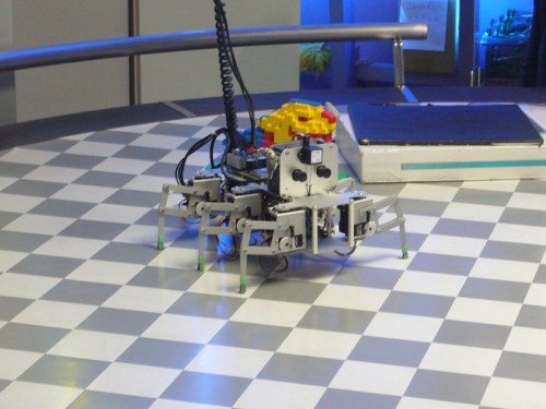 A small robotic spider with multiple legs and sensors, navigating a checkered floor in a tech environment.