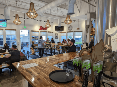 A lively brewery interior with wooden tables, patrons enjoying drinks, and large windows letting in natural light.