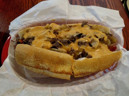 A loaded sandwich with sliced beef, melted cheese, and toppings, served in a toasted hoagie roll on a paper-lined plate.