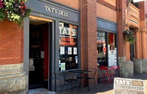 Exterior of Tat's Deli, featuring a brick facade, outdoor seating, and signage for East Coast sandwiches and subs.