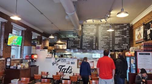 Interior of a casual eatery with a menu board, customers ordering, and a TV displaying a sports game.