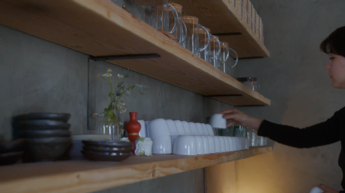 A person arranges white cups on a wooden shelf, surrounded by glassware and decorative items.