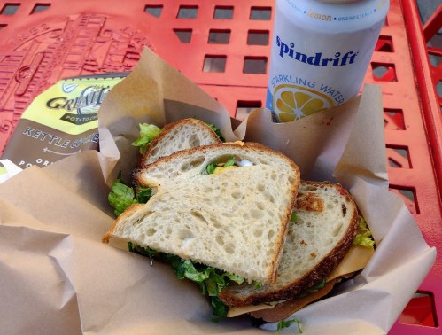 A sandwich with lettuce and cheese on whole grain bread, accompanied by a can of Spindrift sparkling water on a red table.