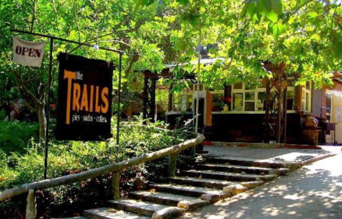 A cozy outdoor café named "The Trails," surrounded by greenery, with a sign indicating it's open.