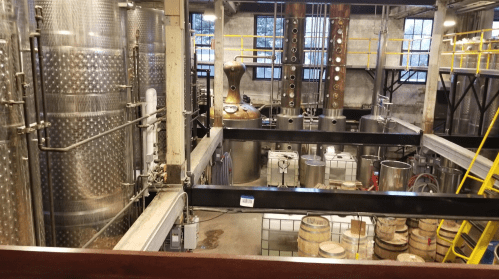 A distillery interior featuring large metal tanks, a copper still, and wooden barrels in a spacious industrial setting.