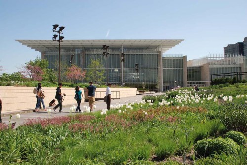 A modern building surrounded by greenery and flowers, with people walking in the foreground on a sunny day.