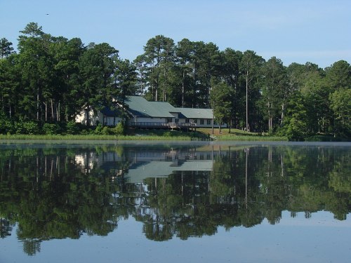 A serene lake reflects a house surrounded by tall trees under a clear blue sky.