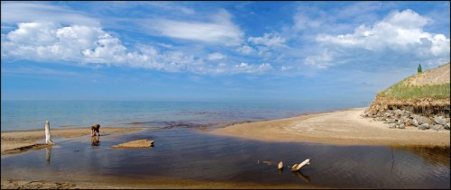 A serene beach scene with a calm sea, sandy shore, and a person near the water under a blue sky with clouds.