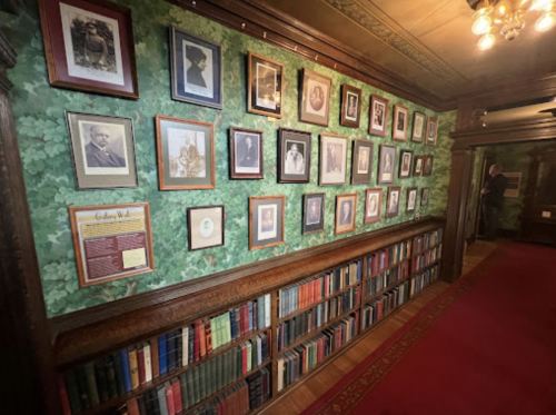 A wall adorned with framed portraits and a bookshelf, set against a green leafy wallpaper in a historic room.