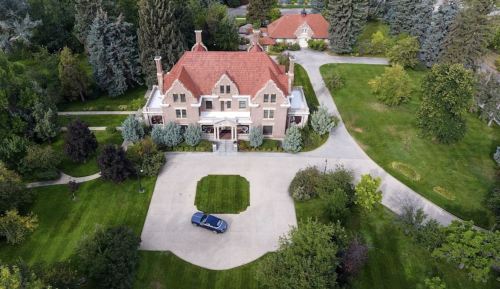 Aerial view of a large, elegant brick mansion with a red roof, surrounded by lush greenery and a circular driveway.