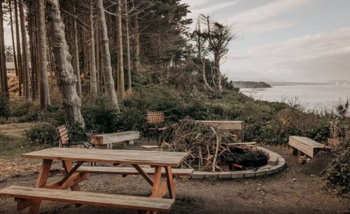 A serene outdoor setting with a picnic table, fire pit, and surrounding trees near a calm body of water.
