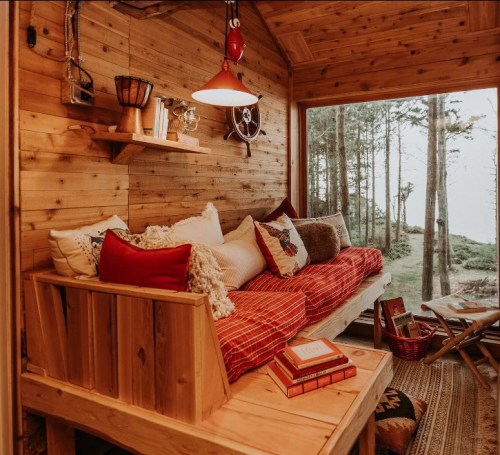 Cozy wooden cabin interior with a red and white sofa, books, and a view of trees outside. Warm, inviting atmosphere.