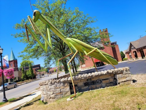 A large green praying mantis sculpture stands on a stone base near a road, with trees and buildings in the background.