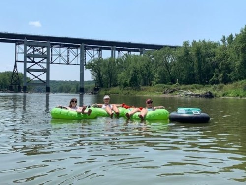 Three people relax on green inner tubes in a river, with a bridge and trees in the background on a sunny day.
