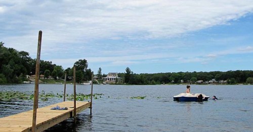 A wooden dock extends over a calm lake, with a person on a float and another swimming nearby, surrounded by greenery.