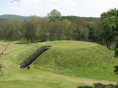 A grassy mound with a wooden walkway, surrounded by trees and a serene landscape under a clear sky.