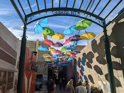 Colorful umbrellas hang overhead in a vibrant alleyway, casting playful shadows on the ground below.