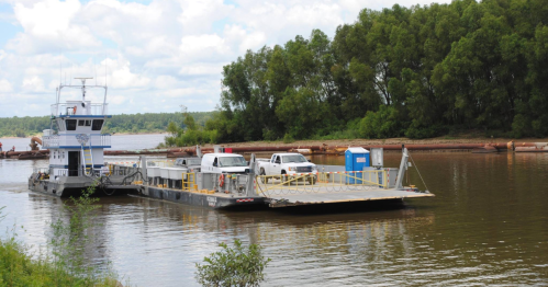 A ferry boat with vehicles on board crosses a river, surrounded by green trees and a cloudy sky.