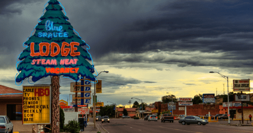 A roadside view featuring a neon sign for the Blue Spruce Lodge, with dark clouds overhead and various businesses nearby.