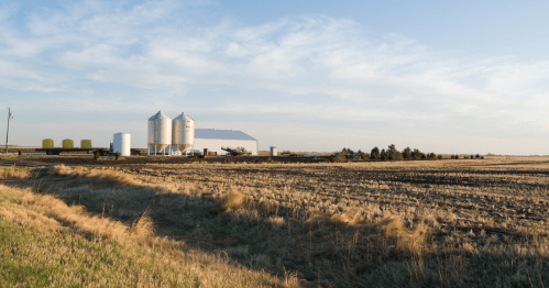 A rural landscape featuring silos and a barn beside a field under a clear blue sky.