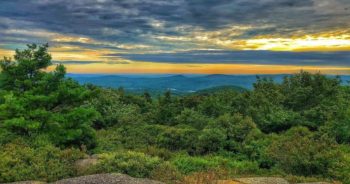 A scenic view of rolling hills and trees under a colorful sunset sky, with clouds scattered above.