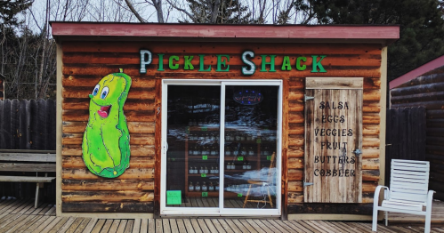 A wooden building with a large pickle mural, featuring a sign that reads "Pickle Shack" and a menu of items for sale.