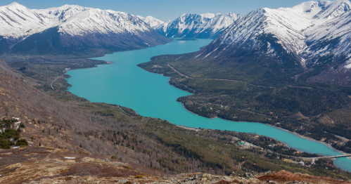 Aerial view of a turquoise river winding through snow-capped mountains and lush green valleys under a clear blue sky.