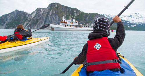 Two kayakers paddle in calm waters near a large ship, surrounded by mountains and snow-capped peaks.