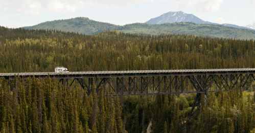 A vehicle crosses a tall, metal bridge surrounded by dense forests and mountains under a partly cloudy sky.