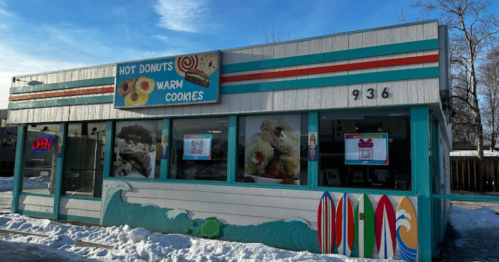 A colorful storefront with a sign for "Hot Donuts Warm Cookies," featuring surfboards and snow in front.
